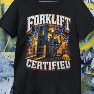 以下が含まれることがあります： 黒いTシャツ。黄色と青のフォークリフトのグラフィックと「FORKLIFT CERTIFIED」の文字。デザインには稲妻とヘルメットが含まれており、文字はかすれたフォントです。