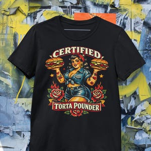 Puede incluir: Camiseta negra con una ilustración de estilo retro. El diseño presenta a una mujer sosteniendo dos hamburguesas, rodeada de rosas y llamas, con el texto "CERTIFIED TORTA POUNDER".