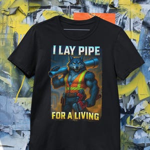 May include: Black t-shirt featuring a muscular wolf-like character in a construction vest, holding a pipe and wrench. Text on the shirt reads "I LAY PIPE FOR A LIVING."