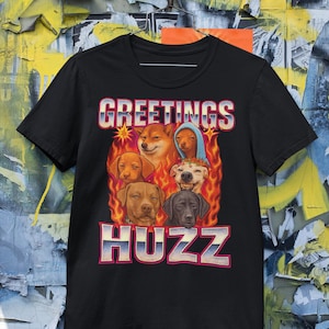 Op de afbeelding: Zwart T-shirt met een afbeelding van verschillende honden gezichten omringd door vlammen. De tekst "GREETINGS HUZZ" staat in een retro lettertype boven en onder de honden. Het shirt heeft korte mouwen.