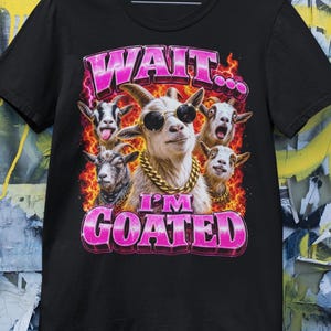 Può includere: T-shirt nero con grafica di cinque capre, una con occhiali da sole e una catena d'oro. Il testo "WAIT... I'M GOATED" è in lettere rosa a bolle con contorno bianco. Lo sfondo è arancione acceso.