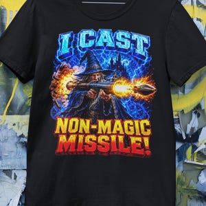 Puede incluir: Una camiseta negra de manga corta con un gráfico de un mago disparando un lanzacohetes. El texto dice I CAST NON-MAGIC MISSILE! El diseño incluye rayos azules, fuego y la silueta de un castillo con letras de color azul y amarillo.