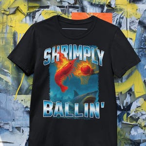 May include: Black t-shirt with a graphic featuring a red shrimp dunking a basketball into a hoop, with a shark below. The text "SHRIMPLY BALLIN'" is in blue and white. The shirt is displayed against a colorful background.