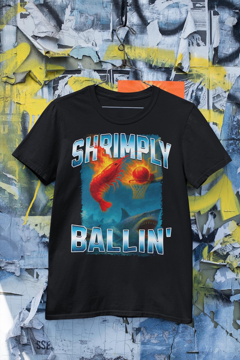 May include: Black t-shirt with a graphic featuring a red shrimp dunking a basketball into a hoop, with a shark below. The text "SHRIMPLY BALLIN'" is in blue and white. The shirt is displayed against a colorful background.