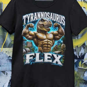 Pode incluir: T-shirt preto com um design de um Tyrannosaurus Rex musculoso a flexionar os bíceps. O design inclui o texto "TYRANNOSAURUS FLEX" em uma fonte estilizada, com detalhes de raios. A camisa também tem o texto "LIFT CRUSH EXTINCT".