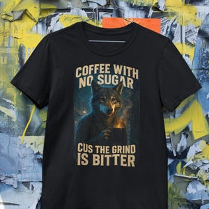 May include: Black t-shirt with a wolf illustration holding a coffee mug. The text on the shirt reads "COFFEE WITH NO SUGAR CUS THE GRIND IS BITTER". The wolf illustration is in shades of gray and brown.