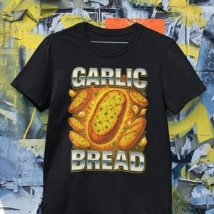 Könnte beinhalten: Schwarzes T-Shirt mit einem Motiv von Knoblauchbrot. Das Design zeigt die Worte "GARLIC BREAD" in Silber und Blau, mit einer Illustration eines goldbraunen Laibs, umgeben von Knoblauchzehen.