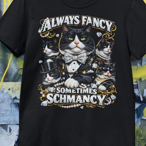 Op de afbeelding: Zwart T-shirt met een afbeelding van katten in smokings, parels en diamanten. De tekst luidt "ALWAYS FANCY SOMETIMES SCHMANCY" in gouden letters.