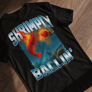 May include: Black t-shirt with a graphic design featuring a red shrimp on fire, a basketball, and a shark. The text "SHRIMPLY BALLIN'" is in a blue and white font. The shirt is displayed on a wooden surface.