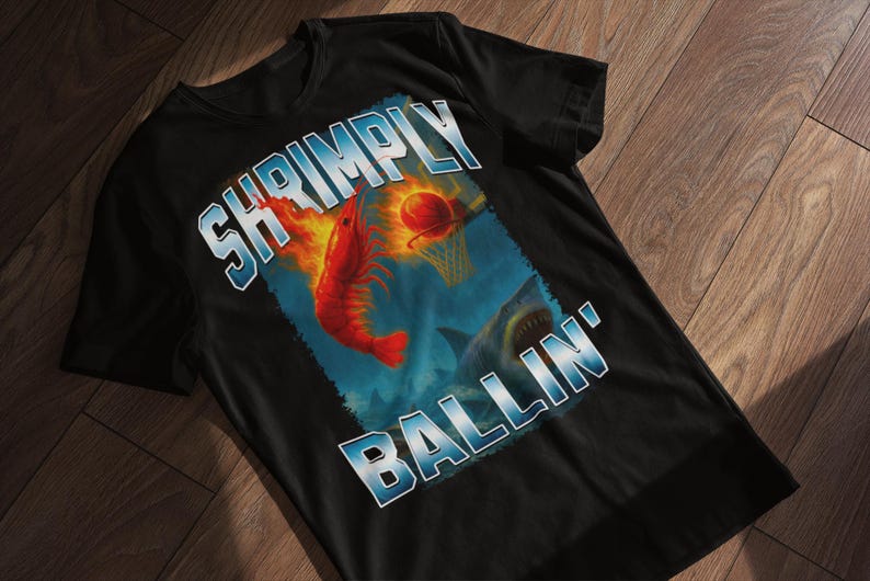 May include: Black t-shirt with a graphic design featuring a red shrimp on fire, a basketball, and a shark. The text "SHRIMPLY BALLIN'" is in a blue and white font. The shirt is displayed on a wooden surface.