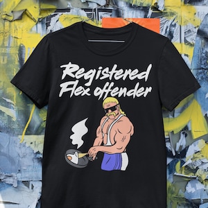 May include: Black t-shirt with the text "Registered Flex Offender" in white script. The shirt features a cartoon illustration of a muscular man with sunglasses cooking an egg in a pan. The man is wearing a blue apron.