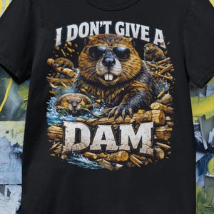 May include: Black t-shirt featuring a beaver wearing sunglasses, with the text "I DON'T GIVE A DAM". The beaver is surrounded by water and logs. A fun, humorous design.