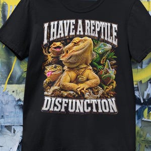 Könnte beinhalten: Schwarzes T-Shirt mit einer Reptilien-Grafik. Das Design zeigt einen Bartagamen, einen Leguan und einen Gecko. Der weiße Text lautet "I HAVE A REPTILE DISFUNCTION" über und unter der Reptilien-Illustration.