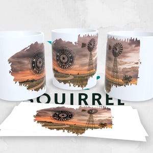 May include: White ceramic mugs with a landscape design featuring a windmill, a sunset, and the text "MINNESOTA WINDESPIRE". The design is printed on the mugs and a matching print on a stack of paper.