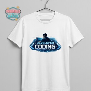 Pode incluir: Camiseta branca com um gr&aacute;fico azul e branco de uma pessoa usando fones de ouvido e trabalhando em um laptop. O texto "DEVELOPER CODING" &eacute; exibido em um design azul estilizado. A camiseta est&aacute; pendurada em um cabide preto.