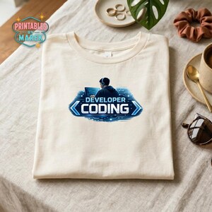 Pode incluir: Uma camiseta creme com um gr&aacute;fico azul e branco que diz "DEVELOPER CODING". O gr&aacute;fico mostra uma pessoa com fones de ouvido trabalhando em um laptop. A camiseta est&aacute; dobrada cuidadosamente sobre uma superf&iacute;cie clara.