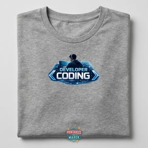 Pode incluir: Camiseta cinza mesclada com um gr&aacute;fico de uma pessoa com fones de ouvido codificando em um laptop. O design inclui as palavras "DEVELOPER CODING" em azul, em um estilo futurista. A camiseta tem decote redondo.