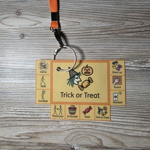 Op de afbeelding: Een oranje keycord bevestigd aan een rechthoekige communicatiekaart met de tekst Trick or Treat. De kaart toont diverse iconen, waaronder een heks, pompoen, snoep en labels zoals Home, Walk, Goodbye, Dress up, Knock, Doorbell, Thank you, In Bucket, Treat en Trick or Treat.