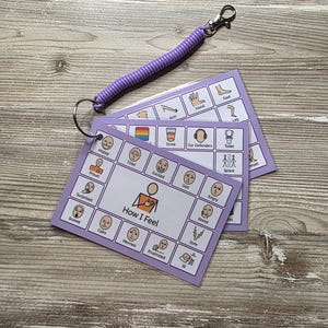 May include: A set of laminated communication cards on a metal ring with a purple coiled wrist strap. The cards feature grid layouts with illustrations and text labels for emotions like Happy, Tired, and Excited, and needs like Drink and Toilet.