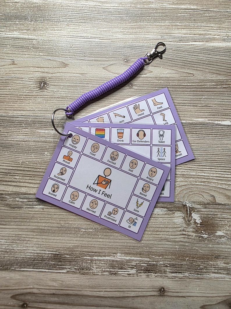 May include: A set of laminated communication cards on a metal ring with a purple coiled wrist strap. The cards feature grid layouts with illustrations and text labels for emotions like Happy, Tired, and Excited, and needs like Drink and Toilet.