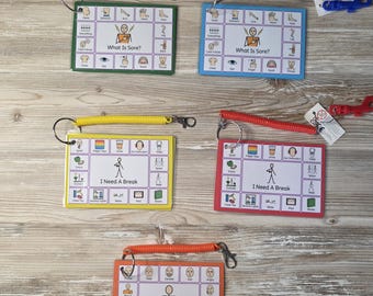 Puede incluir: Cinco tarjetas de comunicación con cordones en espiral y clips. Las tarjetas son verdes, azules, amarillas, rojas y naranjas. Cada tarjeta tiene un título diferente: "What Is Sore?", "I Need A Break" y "How I Feel". Las tarjetas presentan iconos y texto.