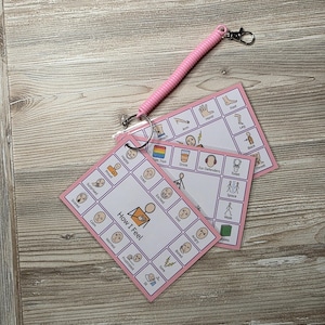 May include: A set of two rectangular communication cards with pink borders attached to a pink coiled wristband with a metal lobster clasp. The cards feature grid layouts with various icons and text labels including How I Feel, Happy, Proud, Surprised, Scared, Calm, Worried, Frustrated, Tired, Excited, Sad, Angry, Bored, Sore, Arm, Hand, Foot, Leg, Back, Drink, Ear Defenders, Toilet, Space, Walk, and Books.
