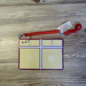 May include: A red and yellow communication board with the words "Now" and "Next" printed on it. The board has a red border and a red coiled lanyard attached to it. The lanyard has a red plastic clip.