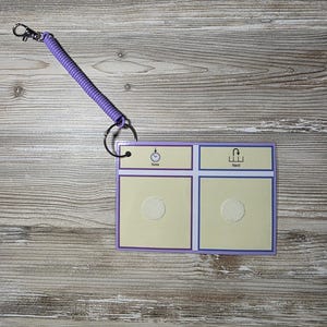 Puede incluir: Una peque&ntilde;a tabla de comunicaci&oacute;n rectangular con un borde morado y un llavero de cord&oacute;n en espiral. La tabla tiene dos secciones etiquetadas como "Ahora" y "Siguiente" con iconos correspondientes. El fondo es de vetas de madera clara.