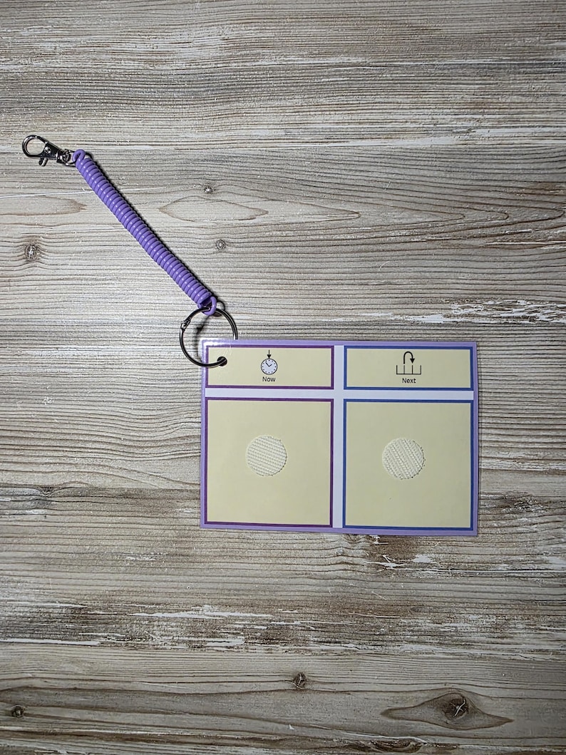 Puede incluir: Una peque&ntilde;a tabla de comunicaci&oacute;n rectangular con un borde morado y un llavero de cord&oacute;n en espiral. La tabla tiene dos secciones etiquetadas como "Ahora" y "Siguiente" con iconos correspondientes. El fondo es de vetas de madera clara.