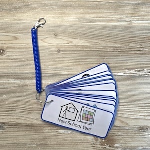 May include: A set of laminated rectangular cards attached to a blue coiled plastic wristband with a metal lobster clasp. The top card features a black line drawing of a person in a house next to a calendar with the text New School Year below.
