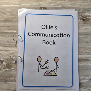 May include: A white communication book bound with two silver metal rings. The cover features a blue rectangular border, the text "Ollie's Communication Book" in black, and a graphic of two stick figures interacting with a colourful tablet.