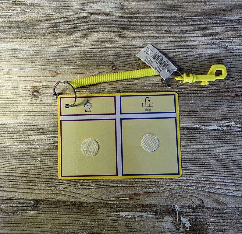 May include: A yellow communication board with a spiral cord and clip. The board has four squares, two with hook-and-loop fasteners. The top of the board has the words "Now" and "Next" with corresponding icons. A tag is attached.