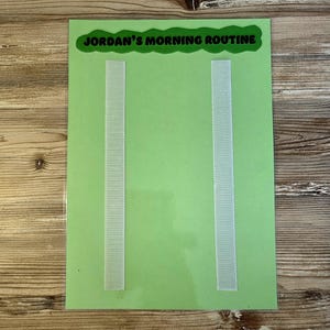 May include: A green chart with the text "Jordan's Morning Routine" at the top. Two vertical strips of white hook-and-loop fasteners are attached to the chart. The chart is on a wooden surface.