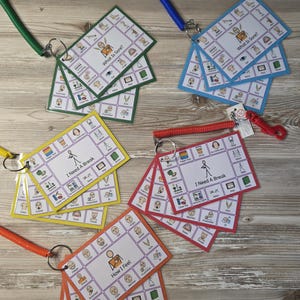 May include: Five sets of communication cards on metal rings with colored plastic coil wristbands. Each set features a central icon and text like What Is Sore, I Need A Break, or How I Feel, surrounded by smaller grid-based icons for daily needs.
