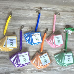 May include: Six sets of communication cards attached to colourful plastic coil wristbands with metal clips. Each set features a square cover card labelled My Communication Cards with an icon, followed by multiple cards in yellow, purple, blue, orange, pink, and green.