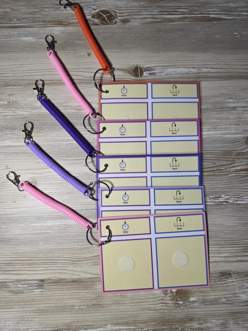 Puede incluir: Un conjunto de tarjetas de comunicaci&oacute;n coloridas con llaveros en espiral. Las tarjetas tienen un fondo amarillo claro con bordes morados y azules. Cada tarjeta tiene las palabras "Now" y "Next". Los llaveros son de color naranja, rosa y morado.