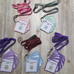 May include: Five sets of My Communication Cards on lanyards in pink, sage green, dark red, light blue, and purple. Each set features a stack of white square cards with black text and a small icon, held together by a metal ring and a coloured plastic border.