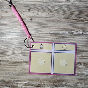 Puede incluir: Un tablero de comunicaci&oacute;n rosa y morado con un cord&oacute;n en espiral y un clip. El tablero tiene dos cuadrados con un c&iacute;rculo texturizado en cada uno, y las palabras "Now" y "Next" encima de los cuadrados. El tablero est&aacute; sujeto a un anillo de metal.