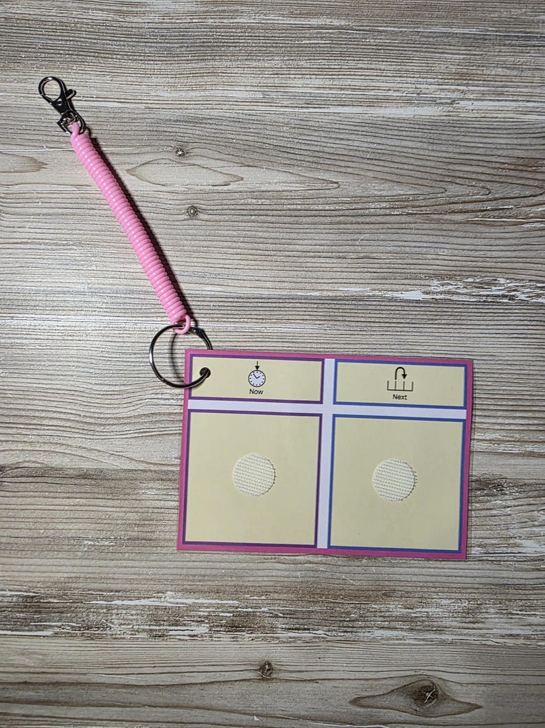 Puede incluir: Un tablero de comunicaci&oacute;n rosa y morado con un cord&oacute;n en espiral y un clip. El tablero tiene dos cuadrados con un c&iacute;rculo texturizado en cada uno, y las palabras "Now" y "Next" encima de los cuadrados. El tablero est&aacute; sujeto a un anillo de metal.