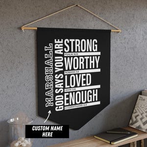 Puede incluir: Un banderín de pared negro con el texto blanco "STRONG, WORTHY, LOVED, ENOUGH". El banderín también incluye el texto "MARSHALL GOD SAYS YOU ARE" y "CUSTOM NAME HERE". Está colgado de una barra de madera.