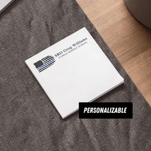 May include: A square white notepad featuring a graphic of a black and white American flag with a thin blue line. The text reads SRO Greg Williams Tupelo Middle School. A black rectangular label with the white text PERSONALIZABLE is visible.