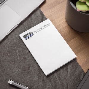 May include: A white rectangular notepad featuring a black and blue thin line flag graphic in the top left corner. The text Sgt. Greg Williams and Owasso Police Department is printed in black ink to the right of the flag graphic on the notepad.