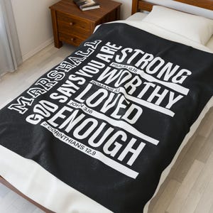 May include: Black blanket with white text that reads "God Says You Are Strong, Worthy, Loved, Enough." The blanket includes Bible verses: Psalm 18:15, Romans 5, John 3:16, and 2 Corinthians 12:9. The blanket is on a bed.