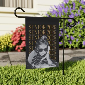 May include: Black garden flag with gold text reading "SENIOR 2026". The flag shows a black and white photo of a child wearing heart-shaped sunglasses and holding a telephone. The flag is displayed on a black metal stand.