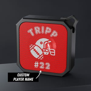 May include: A black portable speaker with a red perforated front grille. The grille features a white graphic of a football helmet and a football, with the text TRIPP arched above and #22 below. A small arrow points to the name with the text CUSTOM PLAYER NAME.