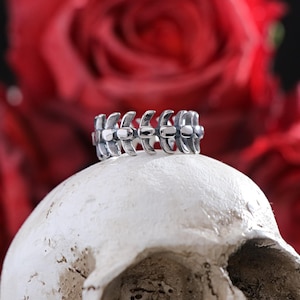 Puede incluir: Un anillo plateado con un diseño repetitivo en forma de hueso, colocado sobre una calavera blanca. El fondo presenta rosas rojas borrosas, creando una estética gótica. El anillo es el punto focal, con detalles visibles.