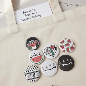 Könnte beinhalten: Eine Sammlung runder Buttons mit verschiedenen Designs, darunter Wassermelonenscheiben, arabischer Text und die palästinensische Flagge. Die Buttons werden auf einer beigen Tragetasche mit einer Karte mit der Aufschrift "Buttons for Palestine - Support & Solidarity" präsentiert.