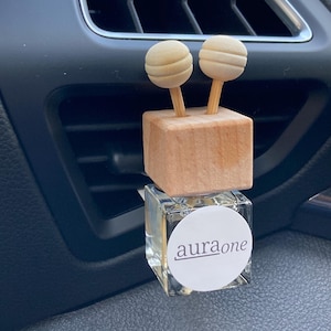 May include: A car air freshener featuring a wooden cube with two spherical wooden toppers. The freshener is mounted on a small, clear glass bottle with a white label reading "auraone". The air freshener is positioned in a car's air vent.