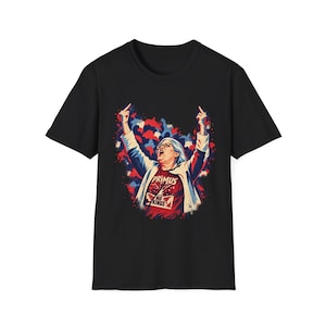 May include: A black short-sleeve t-shirt featuring a graphic of a person with raised arms and middle fingers. The person wears a red shirt with the text "PRIMUS NO KINGS" and a white jacket, set against a red, white, and blue abstract background.
