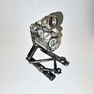 May include: A handcrafted metal sculpture of an engine, constructed from various nuts, bolts, and metal pieces. The sculpture features a cylindrical component and is mounted on a base. The artwork is displayed against a white background.